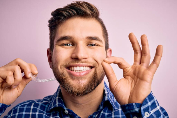 Νεαρός ξανθός άνδρας με γένια και μπλε μάτια κρατώντας οδοντιατρική aligner πάνω από ροζ φόντο κάνει ok υπογράψει με τα δάχτυλα, εξαιρετικό σύμβολο - Φωτογραφία, εικόνα
