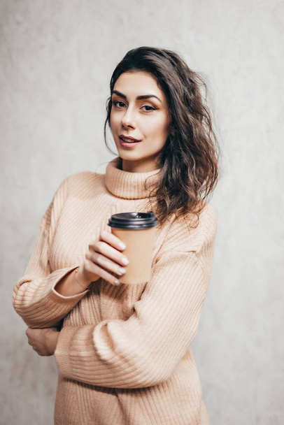 Young beautiful girl in a warm sweater, standing against a wall with a paper cup of coffee. Brunette with a cup of coffee. - Fotó, kép