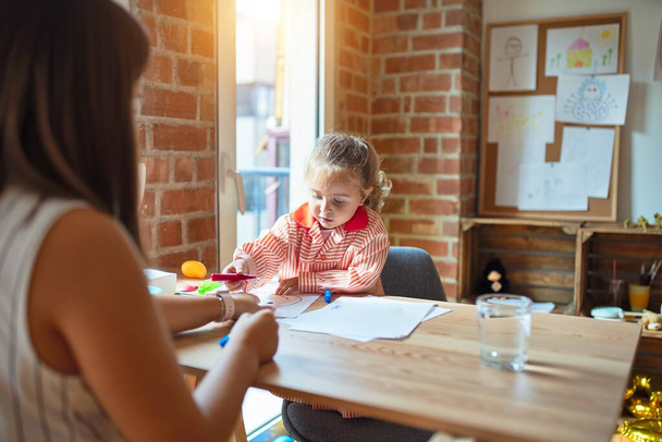 Bella insegnante e studentessa bionda bambina che indossa disegno uniforme scolastica disegnare utilizzando penne colorate all'asilo
 - Foto, immagini