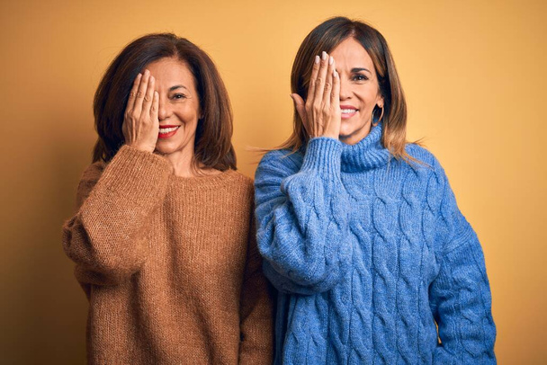 Piękna para sióstr w średnim wieku ubrana w luźny sweter na odizolowanym żółtym tle przykrywająca jedno oko ręką, pewny siebie uśmiech na twarzy i niespodziewane emocje. - Zdjęcie, obraz