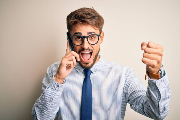 Young businessman having a conversation talking by smartphone over white background screaming proud and celebrating victory and success very excited, cheering emotion - 写真・画像