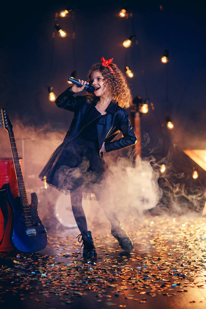 Beautiful girl with curly hair wearing leather jacket, boots sing into a wireless microphone for karaoke like rock star in recording studio or stage. Smoke on background - Foto, imagen