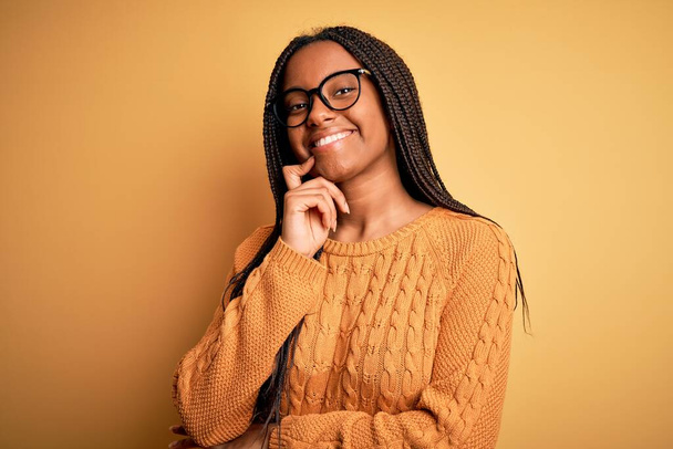 Молодая африканская умная американка в очках и свитере на жёлтом фоне, уверенно смотрящая в камеру со скрещенными руками и поднятой на подбородок рукой. Мысли позитивно
. - Фото, изображение