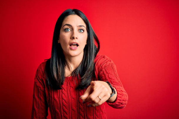 Jeune femme brune aux yeux bleus portant un pull décontracté sur fond rouge isolé pointant mécontent et frustré à la caméra, en colère et furieux contre vous
 - Photo, image