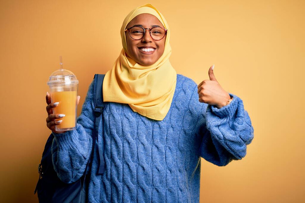 Jovem afro-americana afro turista mulher vestindo hijab muçulmano beber suco de laranja feliz com grande sorriso fazendo sinal ok, polegar com os dedos, excelente sinal
 - Foto, Imagem