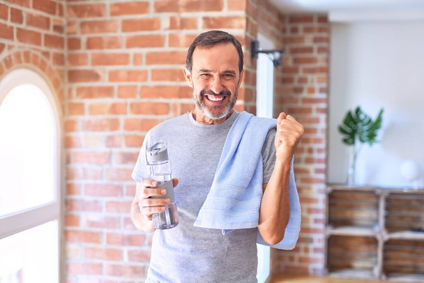 Middle age handsome sportman holding bottle of water and towel standing at gym screaming proud and celebrating victory and success very excited, cheering emotion - Фото, изображение