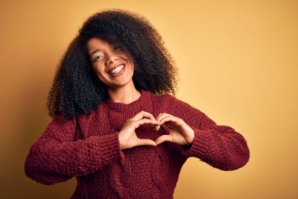 Joven mujer afroamericana hermosa con el pelo afro de pie sobre el fondo aislado amarillo sonriendo en el amor mostrando símbolo del corazón y la forma con las manos. Concepto romántico
. - Foto, imagen