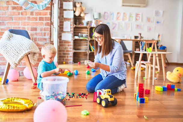 Young caucasian child playing at playschool with teacher. Mother and son at playroom bulding a tower with toy blocks - Foto, imagen