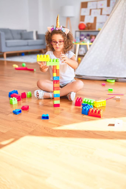 Красивий малюк в окулярах і єдиноріг діадемі сидить, граючи з будівельними блоками в дитячому саду
 - Фото, зображення