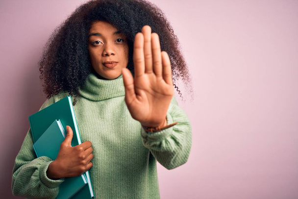 Mladá africká americká studentka s afro vlasy čtení knih přes růžové pozadí s otevřenou rukou dělat stop sign s vážným a sebejistý výraz, obrana gesto - Fotografie, Obrázek