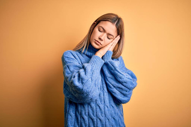 Young beautiful blonde woman wearing turtleneck sweater over yellow isolated background sleeping tired dreaming and posing with hands together while smiling with closed eyes. - Φωτογραφία, εικόνα