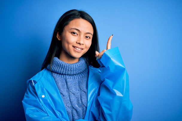 Młoda piękna Chinka ubrana w płaszcz przeciwdeszczowy stojąca nad odizolowanym niebieskim tłem z dużym uśmiechem na twarzy, wskazująca ręką i palcem na bok patrzącą w kamerę. - Zdjęcie, obraz