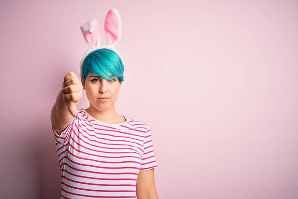 Молодая женщина с модными голубыми волосами, носящая пасхальные кроличьи уши на розовом фоне, выглядит несчастной и злой, показывая отказ и негатив с опущенными пальцами. Плохое выражение
. - Фото, изображение