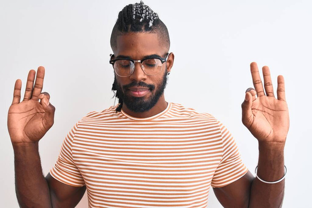 Hombre afroamericano vistiendo camiseta a rayas y gafas sobre fondo blanco aislado relajarse y sonreír con los ojos cerrados haciendo gesto de meditación con los dedos. Concepto de yoga
. - Foto, Imagen