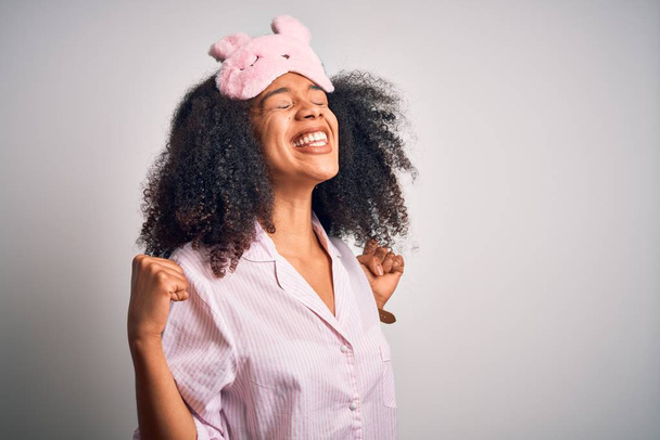 Nuori afrikkalainen amerikkalainen nainen afro hiukset yllään nukkuva silmä naamio ja vaaleanpunainen pyjama erittäin onnellinen ja innoissaan tekee voittaja ele kädet kohotettuna, hymyillen ja huutaen menestystä. Juhlan käsite
. - Valokuva, kuva