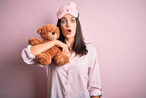Jeune femme brune aux yeux bleus portant un pyjama embrassant un ours en peluche animal empaillé effrayé sous le choc avec un visage surprise, effrayé et excité par l'expression de la peur
 - Photo, image