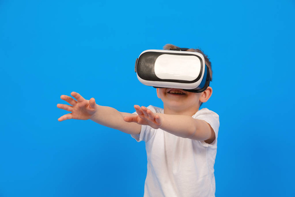 Boy in virtual reality glasses VR holds his hands in front of him and tries to play with virtual world, wearing white t-shirt, denim pants on blue background. Innovative technology, education concept - Zdjęcie, obraz