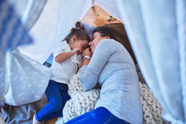 Young beautiful teacher and toddler smiling with happy face lying down over blanket inside tipi at kindergarten - Photo, image