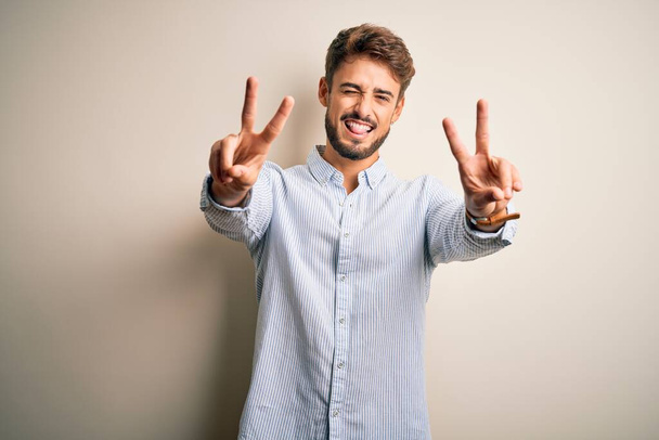 Young handsome man with beard wearing striped shirt standing over white background smiling with tongue out showing fingers of both hands doing victory sign. Number two. - Fotó, kép