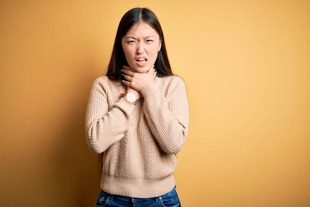 Young beautiful asian woman wearing casual sweater over yellow isolated background shouting suffocate because painful strangle. Health problem. Asphyxiate and suicide concept. - Фото, изображение