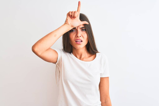 Young beautiful woman wearing casual t-shirt standing over isolated white background making fun of people with fingers on forehead doing loser gesture mocking and insulting. - 写真・画像