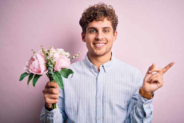 Young blond man with curly hair holding beautiful bouquet over isolated pink background very happy pointing with hand and finger to the side - Foto, imagen