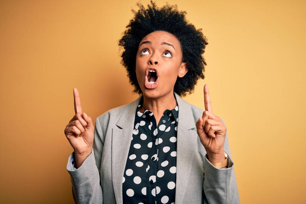 Young beautiful African American afro businesswoman with curly hair wearing jacket amazed and surprised looking up and pointing with fingers and raised arms. - Photo, Image