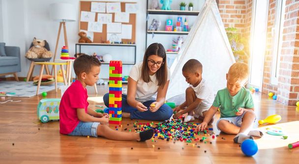 Молодий красивий вчитель і малюки грають з будівельними блоками навколо безлічі іграшок в дитячому саду
 - Фото, зображення