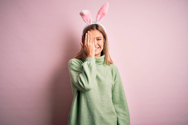 Молода красива жінка в великодніх вухах кролика стоїть над ізольованим рожевим тлом, покриваючи одне око рукою, впевнено посміхається обличчям і дивує емоціями
. - Фото, зображення