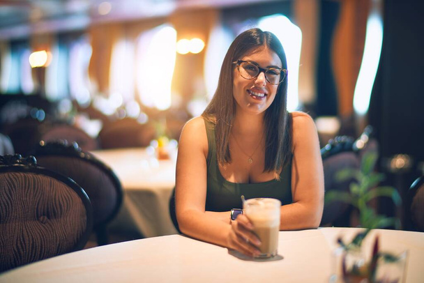 Молодая красивая женщина улыбается счастливой и уверенной. Сидя с улыбкой на лице держа стакан кофе в ресторане
 - Фото, изображение