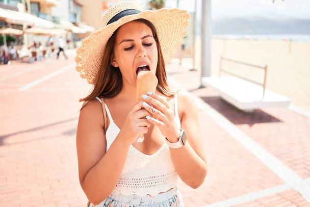 Young beautiful woman eating ice cream cone by the beach on a sunny day of summer on holidays - Photo, Image