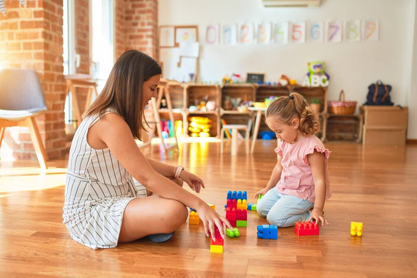 Hermosa maestra y rubia niña construyendo torre usando bloques de plástico en el jardín de infantes
 - Foto, Imagen