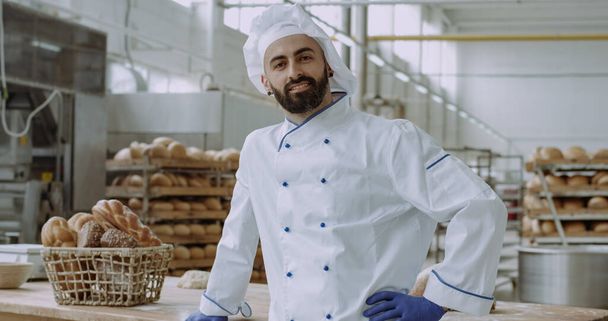 Μπροστά από το πορτραίτο της κάμερας ενός εμφανίσιμου άντρα με εμπορικό πρόσωπο φούρναρη σε μια κομψή στολή χαμογελώντας μεγάλο ενώ στέκεται δίπλα από ένα τραπέζι εργασίας σε μια κουζίνα αρτοποιίας - Φωτογραφία, εικόνα