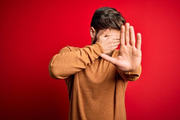 Young blond man with beard and blue eyes wearing casual sweater over red background covering eyes with hands and doing stop gesture with sad and fear expression. Embarrassed and negative concept. - Photo, Image