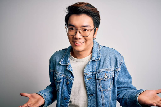 Young handsome chinese man wearing denim jacket and glasses over white background clueless and confused expression with arms and hands raised. Doubt concept. - Foto, immagini