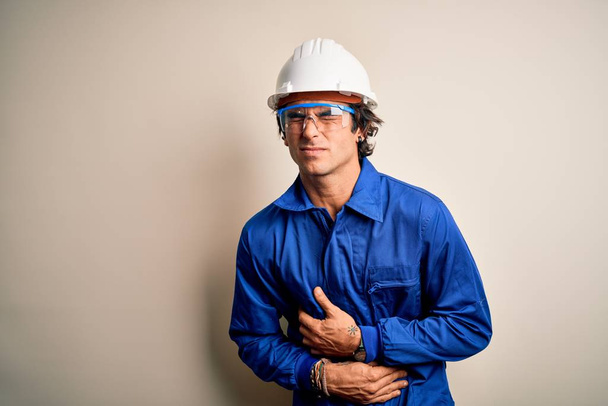 Young constructor man wearing uniform and security helmet over isolated white background with hand on stomach because nausea, painful disease feeling unwell. Ache concept. - Photo, Image