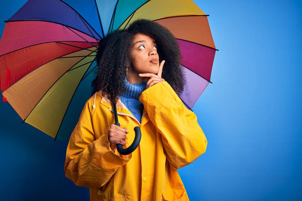 Young african american woman with afro hair under colorful umbrella wearing winter coat for rain serious face thinking about question, very confused idea - Valokuva, kuva