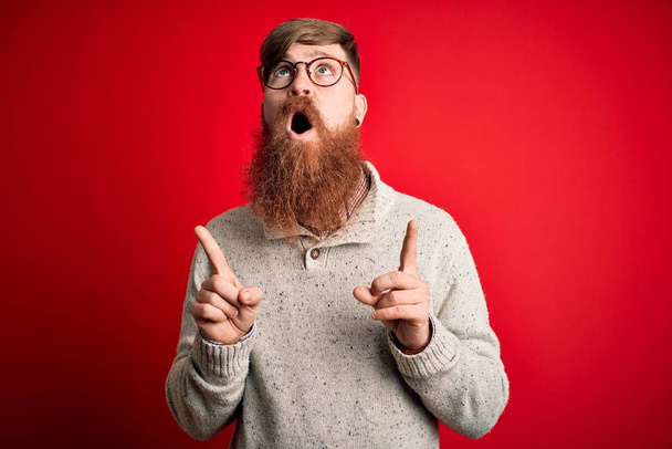 Bello irlandese rossa uomo con barba indossare maglione casual e occhiali su sfondo rosso stupito e sorpreso guardando in alto e puntando con le dita e braccia alzate
. - Foto, immagini