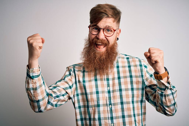 Knappe Ierse roodharige man met baard die een bril draagt en een hippe overhemd dat verrast en verbaasd is voor succes met opgeheven armen en open ogen. Winnaars concept. - Foto, afbeelding