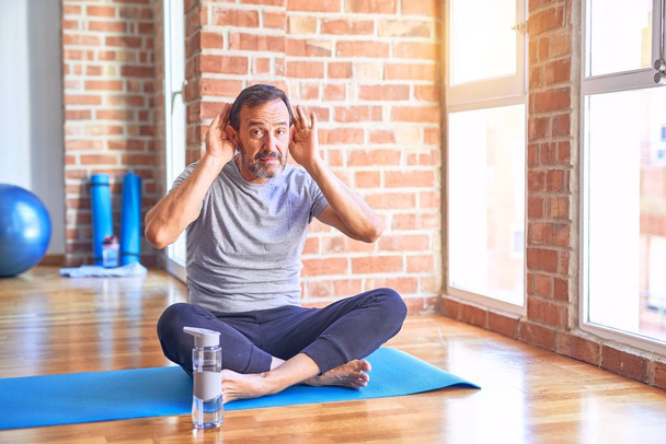 Przystojny sportowiec w średnim wieku, siedzący na macie i ćwiczący jogę na siłowni, próbujący usłyszeć obie ręce na uchu, ciekawy plotek. Problemy ze słuchem, głuchy - Zdjęcie, obraz