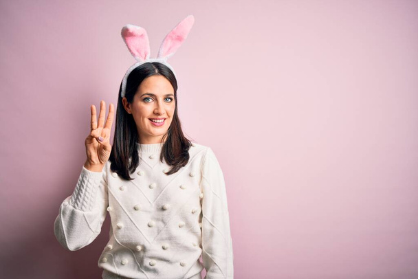 Joven mujer caucásica con lindas orejas de conejo de Pascua sobre fondo rosa aislado mostrando y señalando hacia arriba con los dedos número tres mientras sonríe confiado y feliz
. - Foto, Imagen