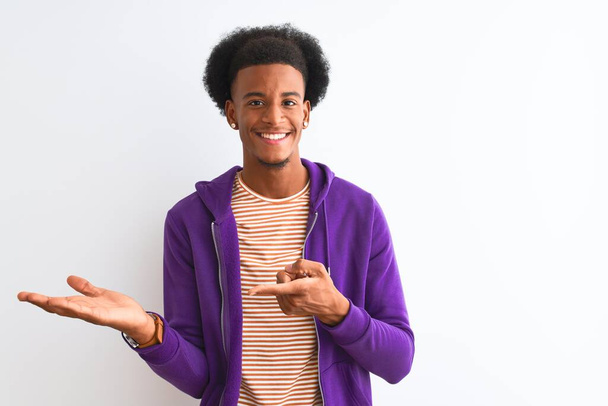 Afrykański Amerykanin ubrany w purpurową bluzę stojący nad odizolowanym białym tłem zdumiony i uśmiechnięty do kamery prezentując się z ręką i wskazując palcem. - Zdjęcie, obraz