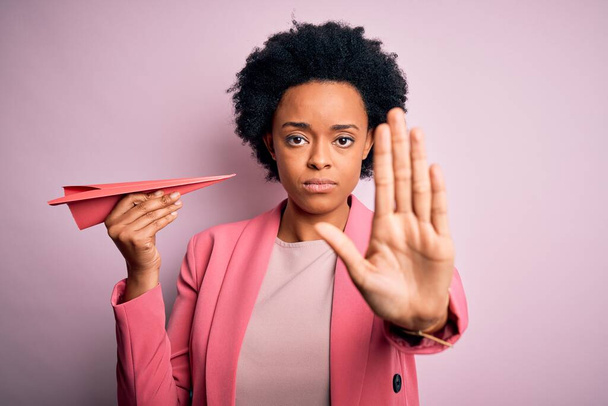 Nuori Afrikkalainen Amerikan afro nainen kihara tukka tilalla vaaleanpunainen paperi lentokone avoimin käsin tekee stop merkki vakava ja luottavainen ilme, puolustus ele
 - Valokuva, kuva