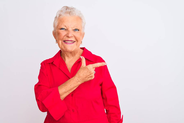 Старша сіро-волохата жінка в червоній повсякденній сорочці стоїть над ізольованим білим тлом весела посмішка спрямована рукою і пальцем в сторону з щасливим і природним виразом на обличчі
 - Фото, зображення
