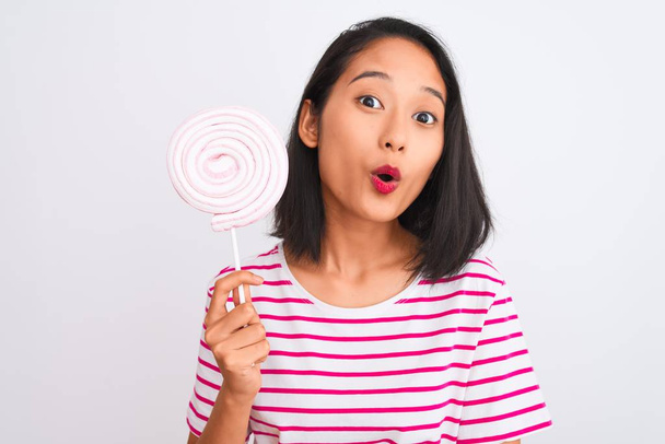 Young beautiful chinese woman eating lollipop standing over isolated white background scared in shock with a surprise face, afraid and excited with fear expression - Photo, Image
