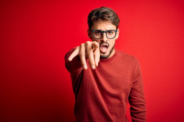 Mladý pohledný muž s vousy nosí brýle a svetr stojící nad červeným pozadím ukazuje nespokojeně a frustrovaný na kameru, naštvaný a vzteklý na vás - Fotografie, Obrázek