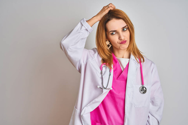 Redhead caucasian doctor woman wearing pink stethoscope over isolated background confuse and wonder about question. Uncertain with doubt, thinking with hand on head. Pensive concept. - Valokuva, kuva