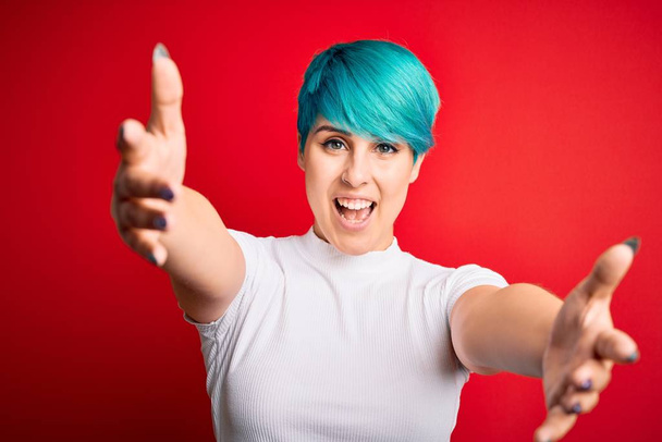 Young beautiful woman with blue fashion hair wearing casual t-shirt over red background looking at the camera smiling with open arms for hug. Cheerful expression embracing happiness. - Фото, изображение
