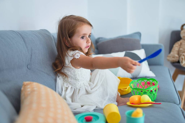 Suloinen blondi lapsi istuu sohvalla ruoanlaitto ruoan ja ruokailuvälineiden leluja kotona
 - Valokuva, kuva