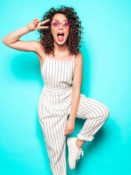 Portrait of beautiful smiling model with afro curls hairstyle dressed in summer hipster clothes.Sexy carefree girl posing in studio near blue wall.Trendy funny and positive woman shows peace sign - Фото, изображение
